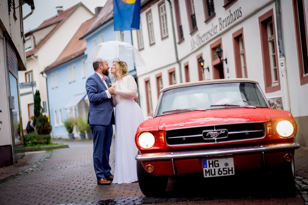 Brautpaar steht gemeinsam neben einem roten Hochzeitsauto